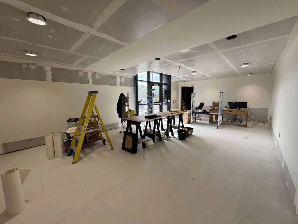 The interior of the new Dundee Design Studio at Riverside House, Luna Place, part way through fit-out, with a boarded and jointed ceiling, painted walls, full height glazing at the entrance, a decorator's ladder and trestle table, and a desk set up at the far end
